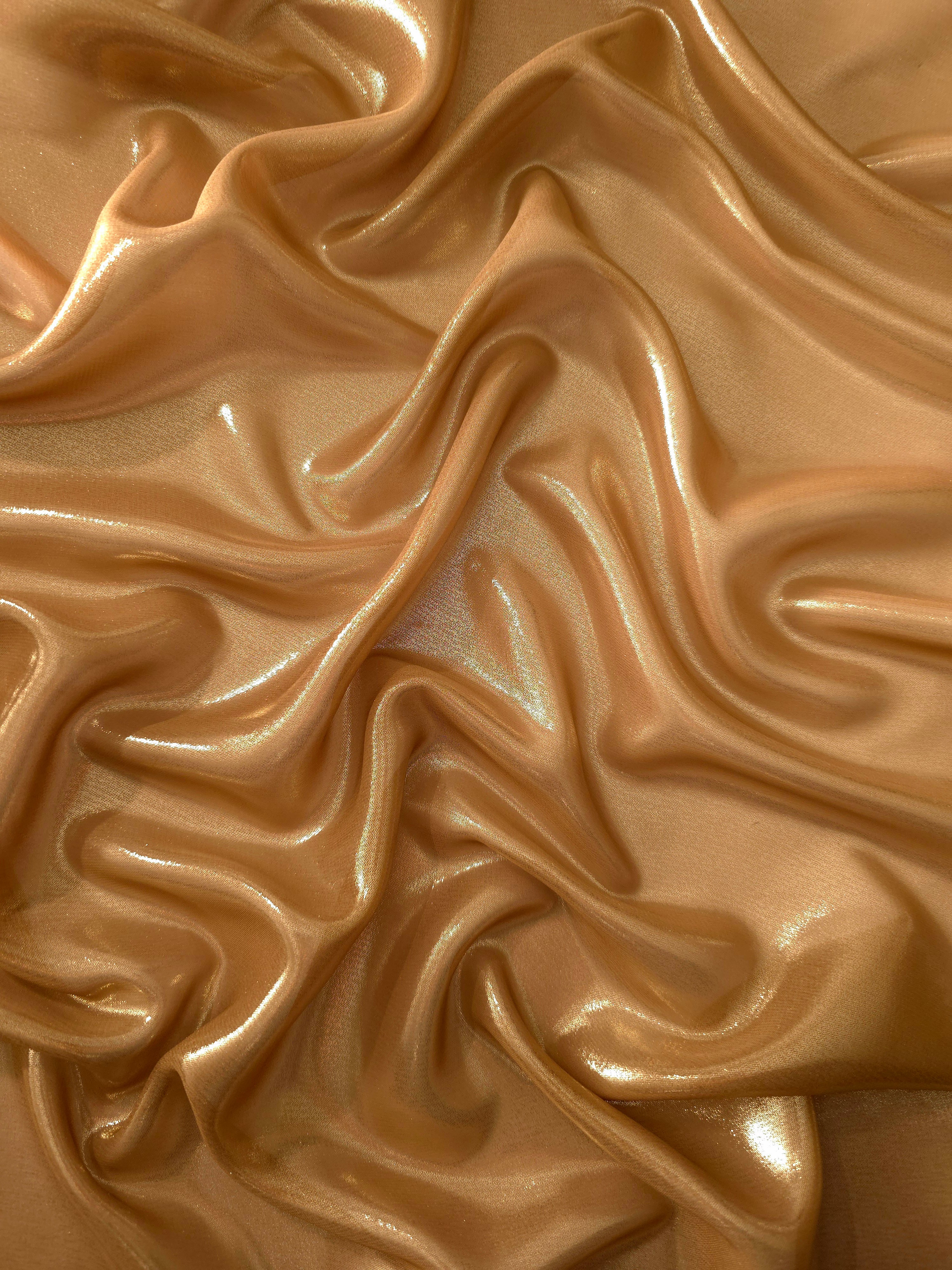 Golden tan surface detail view of liquid metallic shimmer chiffon fabric, highlighting fluid folds and reflective surface