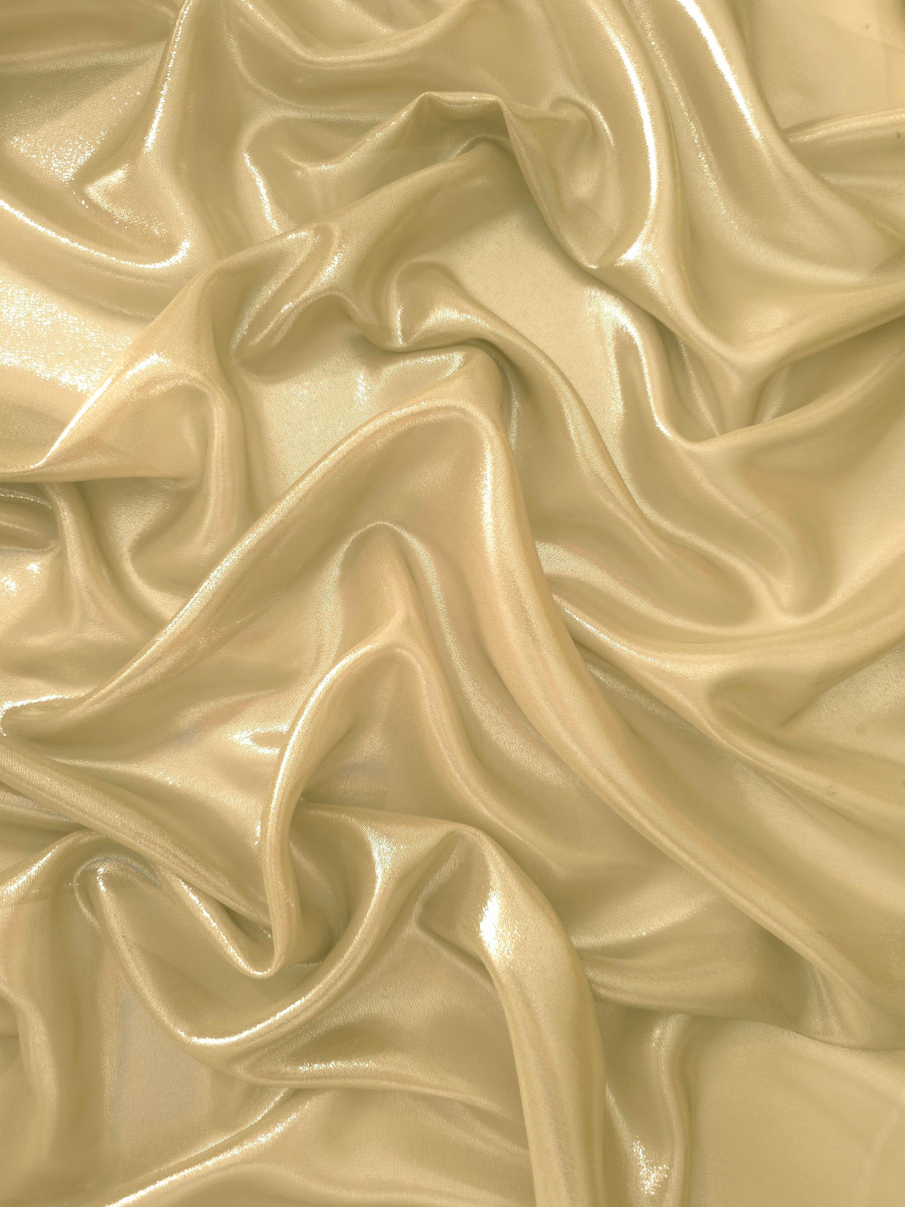 Pale gold surface detail view of liquid metallic shimmer chiffon fabric, highlighting fluid folds and reflective surface